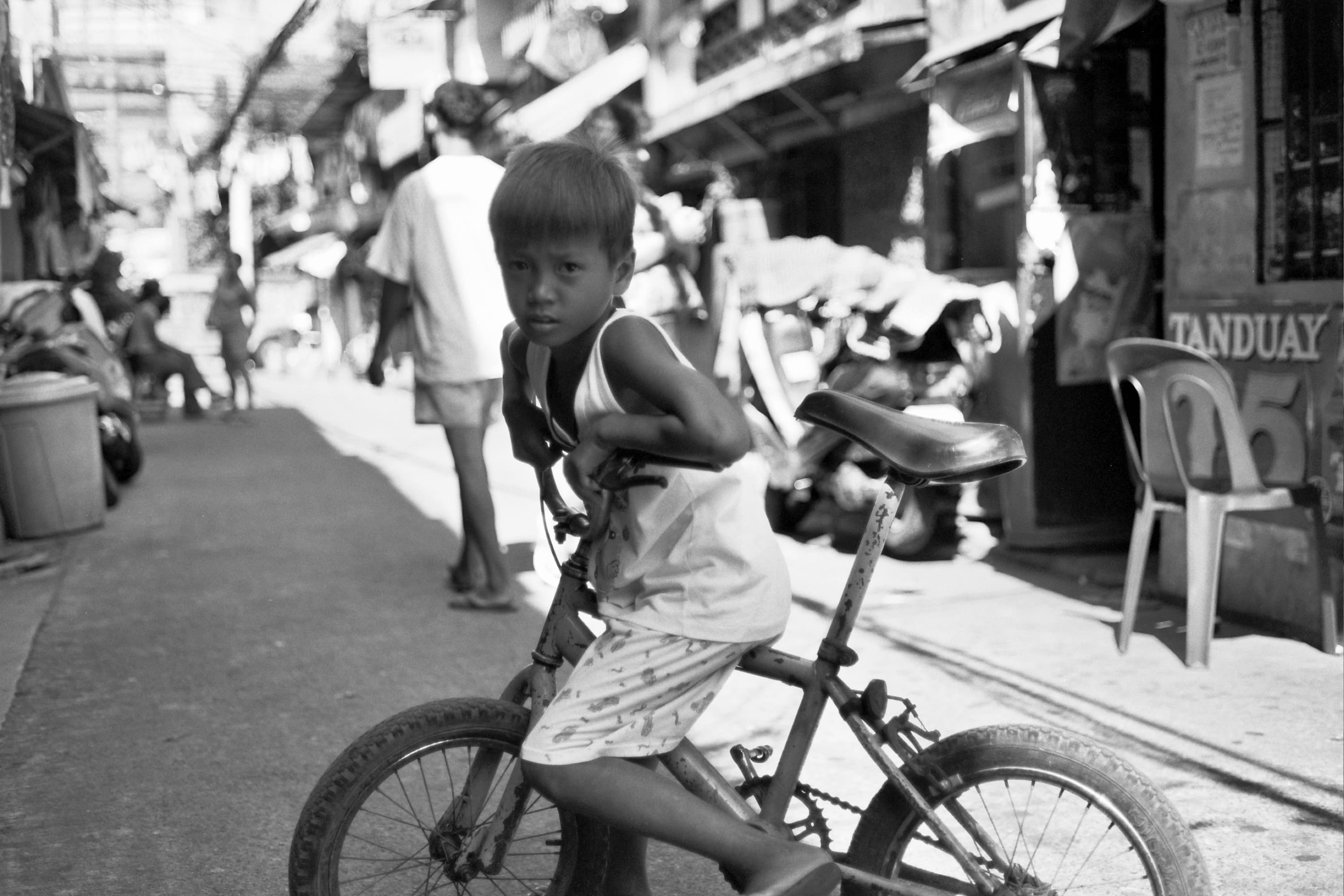 Young boy on a worn bicycle glancing at the camera in a busy street beside a Tanduay sign