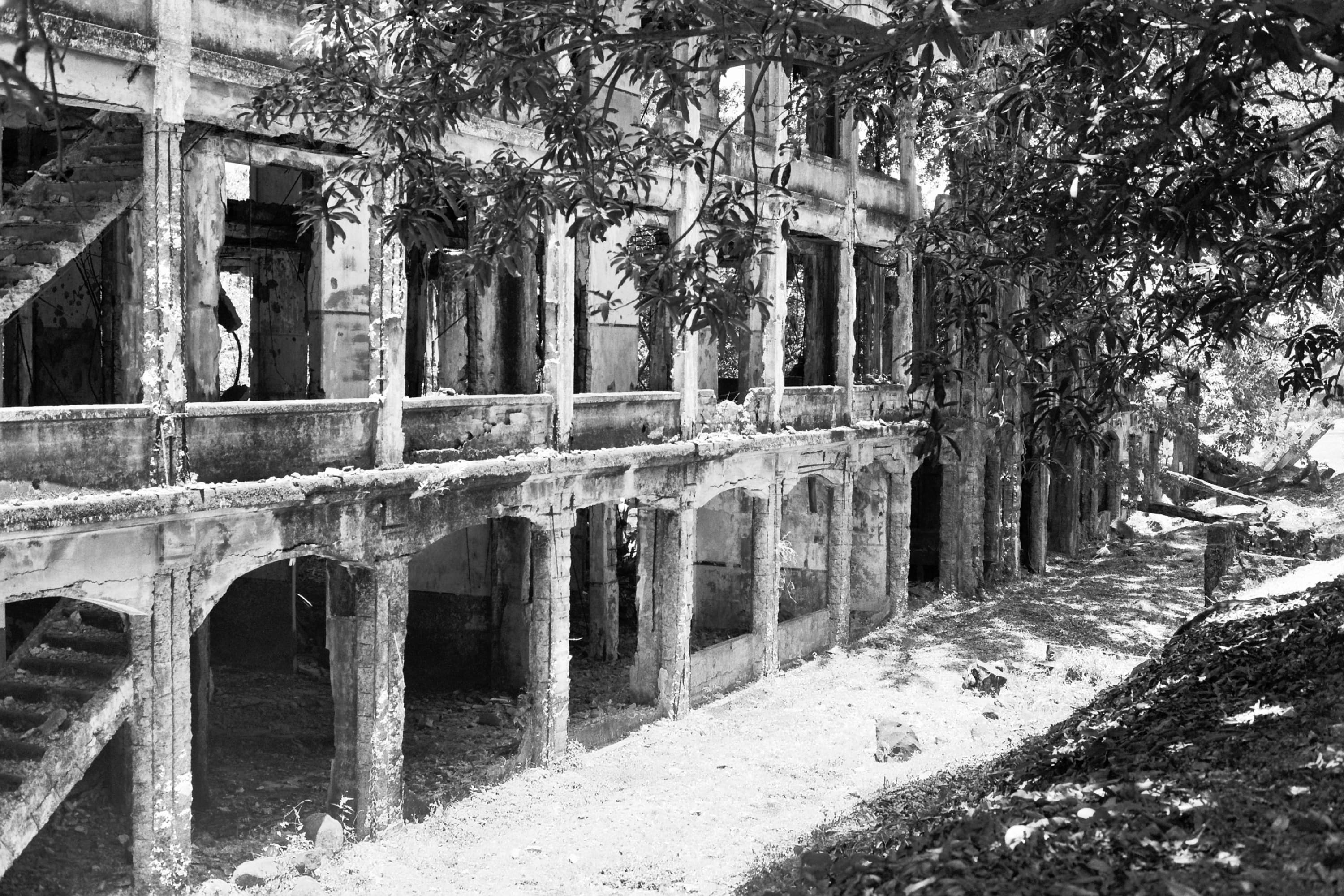 Bombed-out concrete barracks ruins with arches and stairways overgrown by trees