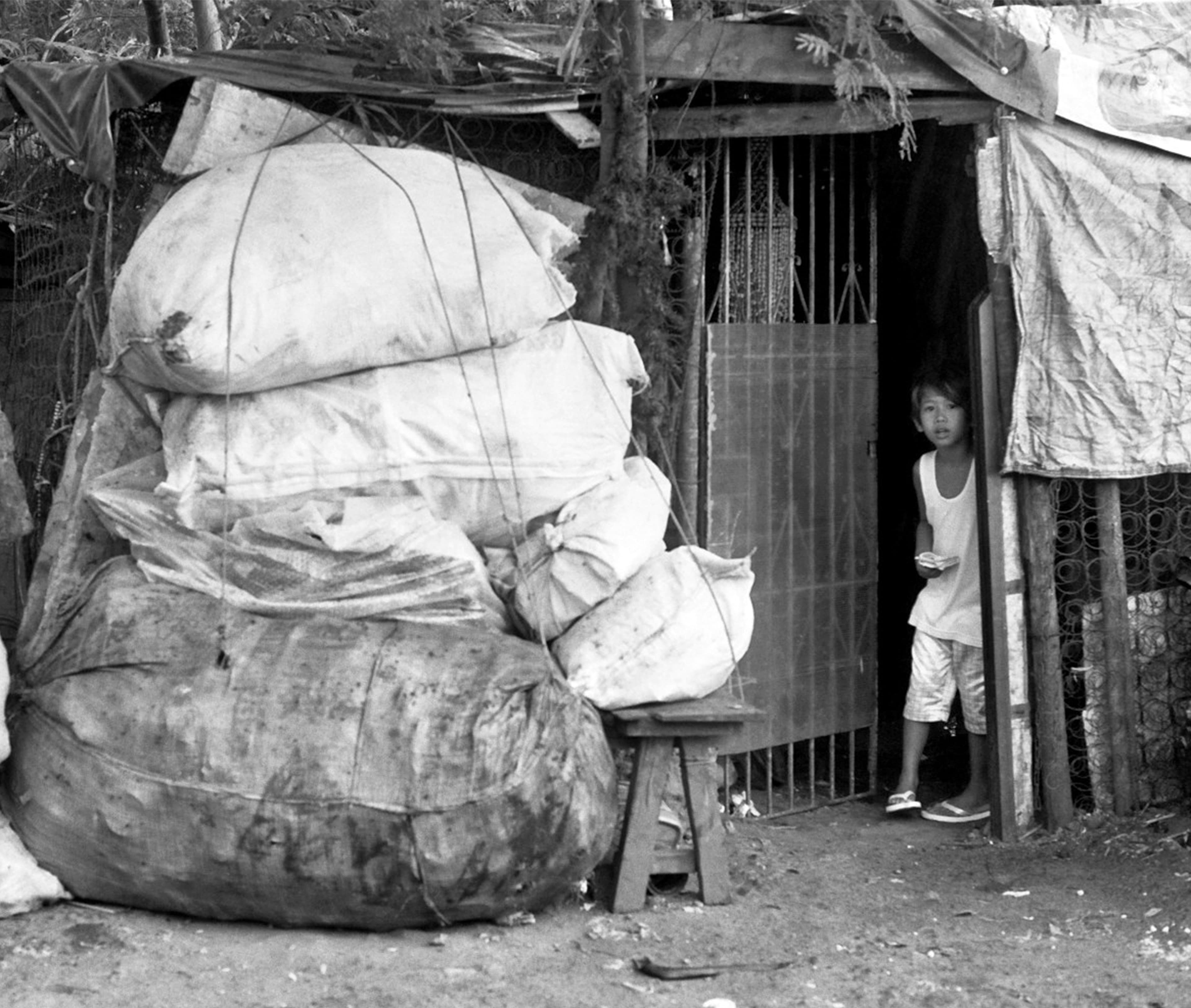 Child peering from the doorway of a makeshift home beside huge tied sacks