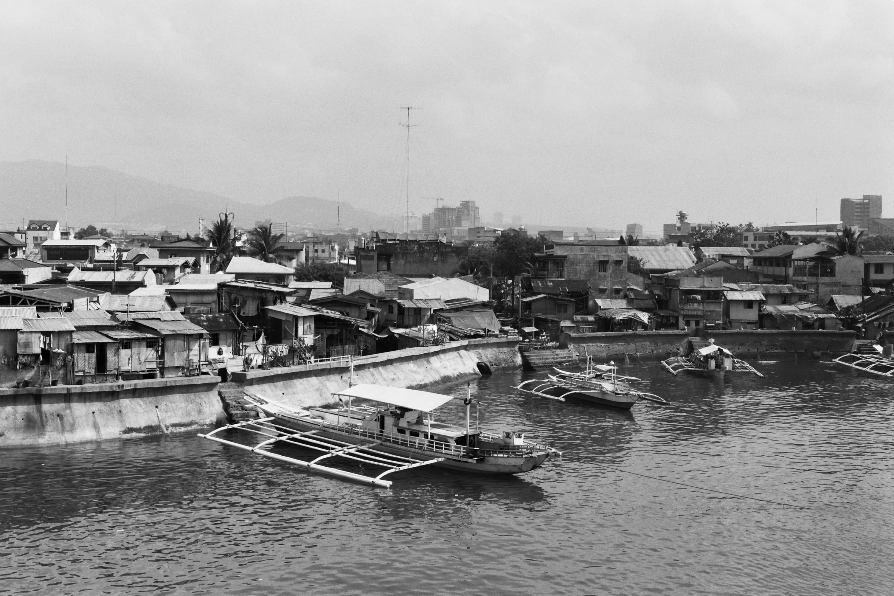 Outrigger boats in a harbor before densely packed tin-roofed houses and hazy mountains