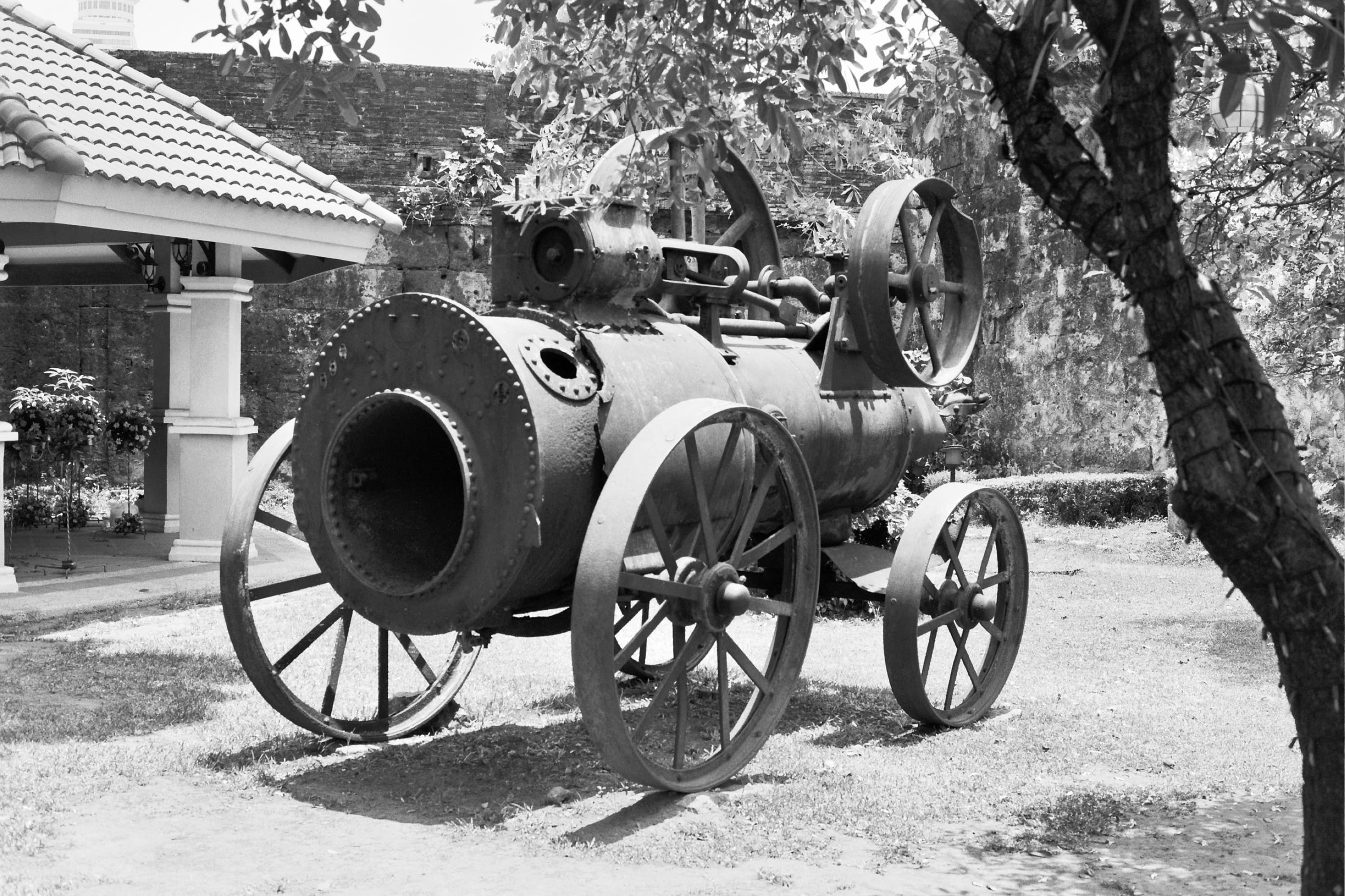 Rusted steam engine with spoked iron wheels beneath a tree against old stone walls