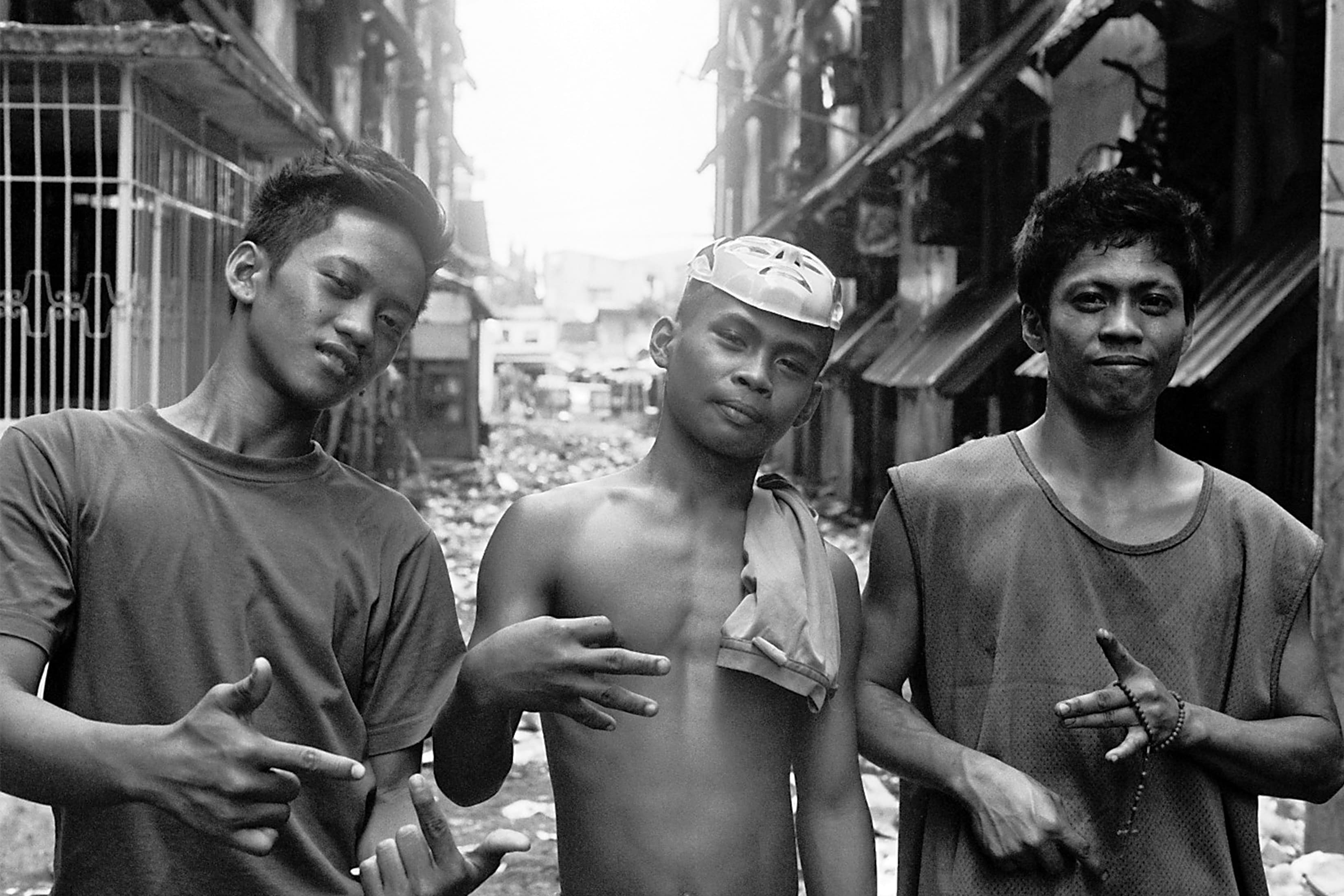 Three young men posing with hand signs on a rubble-strewn Manila street, one wearing a mask on his head