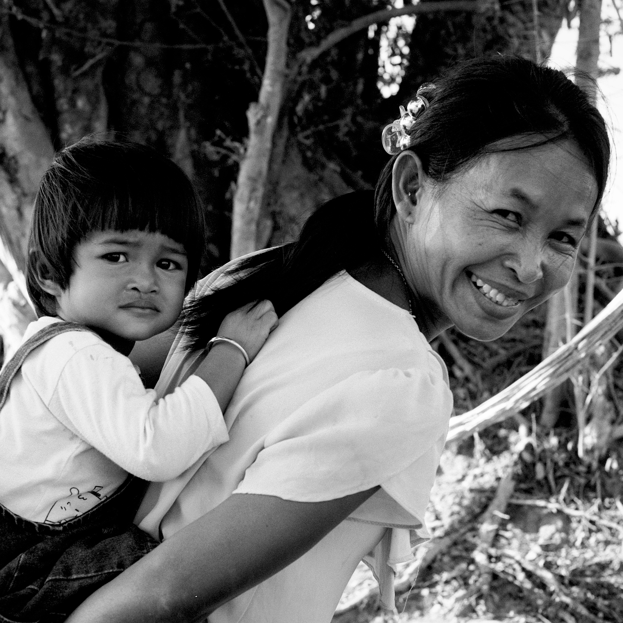 Smiling Cambodian mother carrying young child on her back in natural outdoor setting