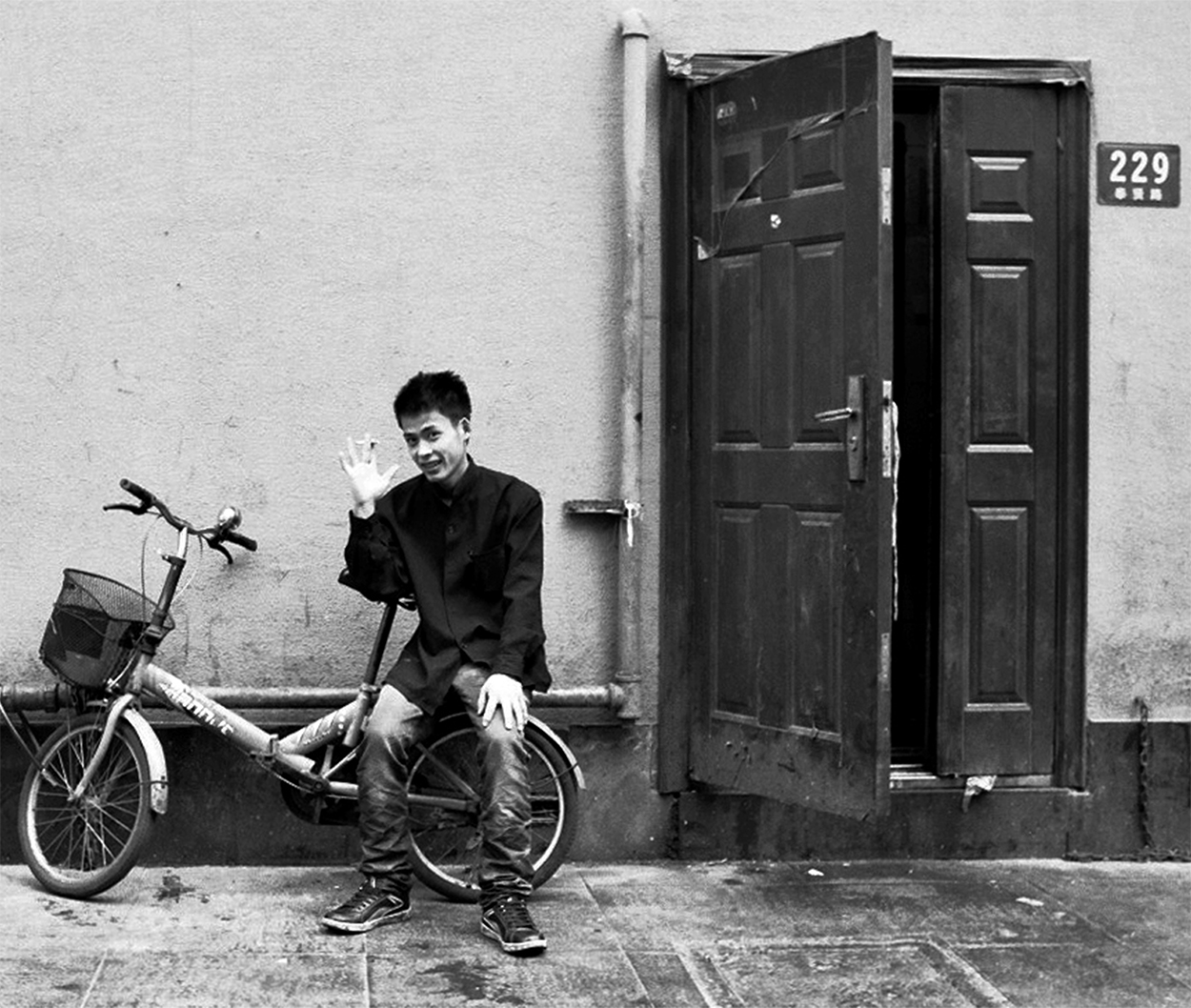 Young man on bicycle waving in front of weathered wall with dark wooden doors in Chinese alley