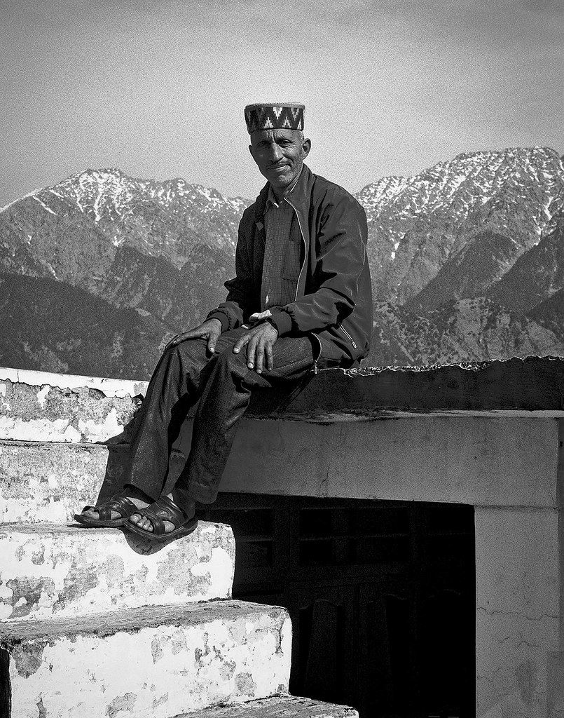 Man in traditional Himachali cap sitting on rooftop with Himalayan mountains