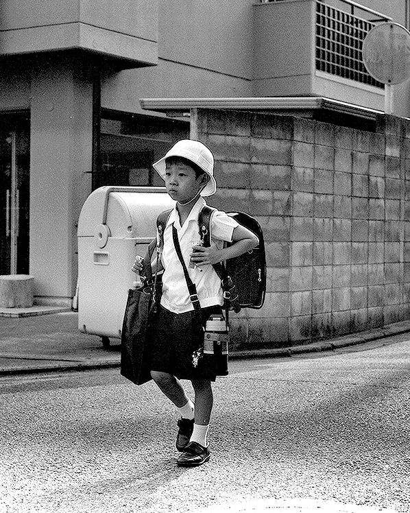 Japanese schoolchild in white helmet and uniform with backpack walking past modern residential architecture