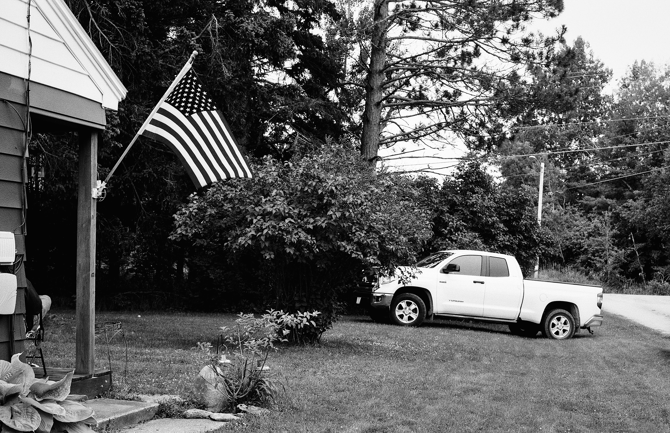 White truck parked in driveway with American flag on pole, white house, and dense forest background